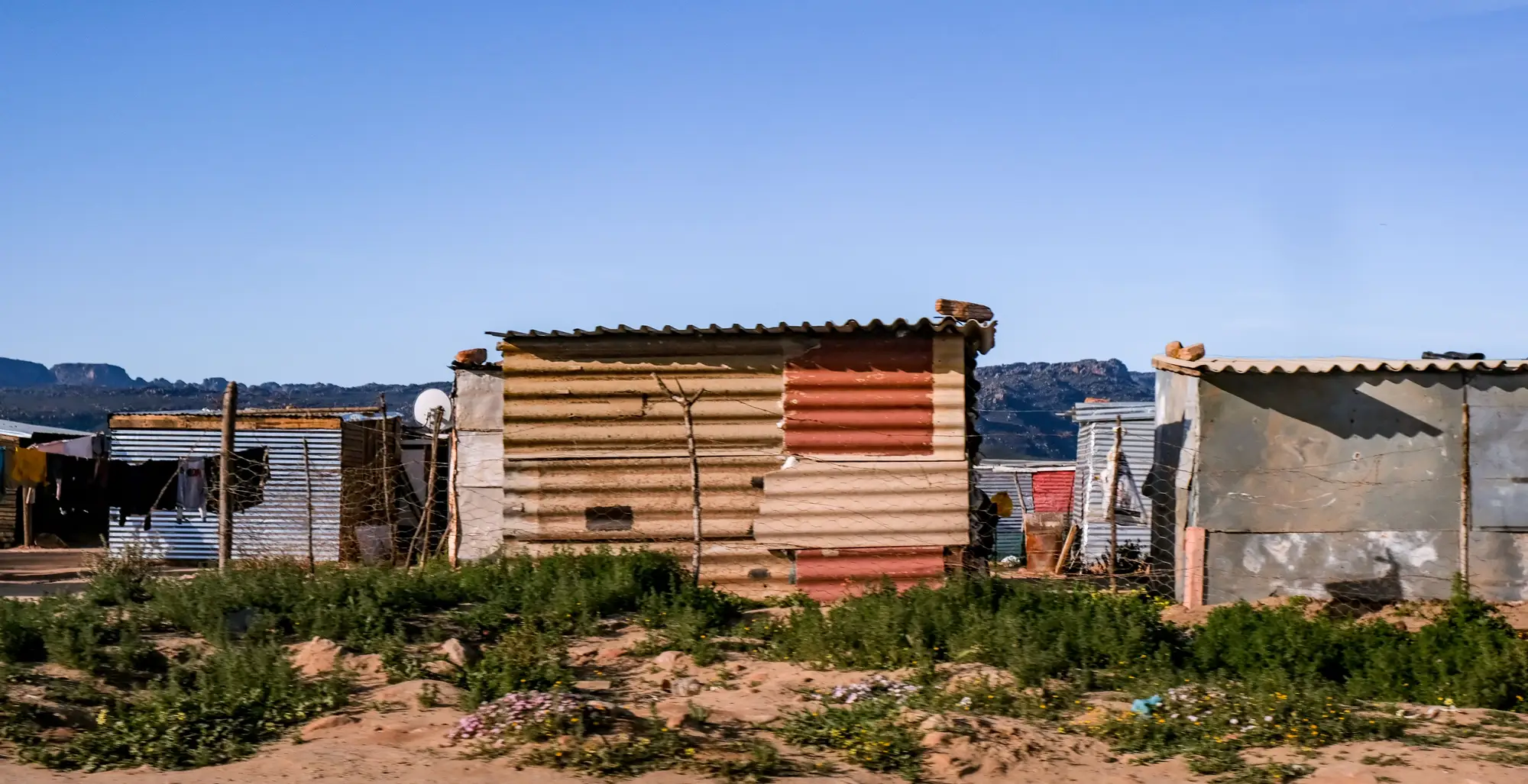Corrugated metal houses on sandy ground under a clear sky, seen from the track