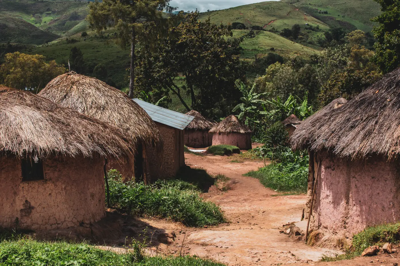 A cluster of thatched-roof huts connected by dirt paths in a rural village, with green hills behind.